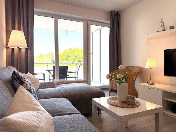 A modern living room with a sofa, a coffee table, a TV, and a balcony with outdoor furniture.
