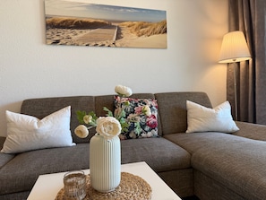 Un salon avec un canapé, des coussins, un vase de fleurs, une lampe et un tableau au mur.