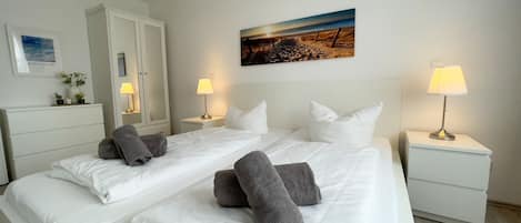 Habitación de hotel con dos camas, ropa de cama blanca, un cuadro de una playa, un armario y lámparas de noche.