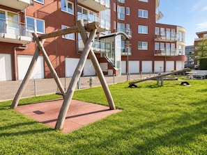 Ein Spielplatz mit Schaukeln und einer Wippe in einer grünen Wiese, im Hintergrund ein Backsteinhaus.