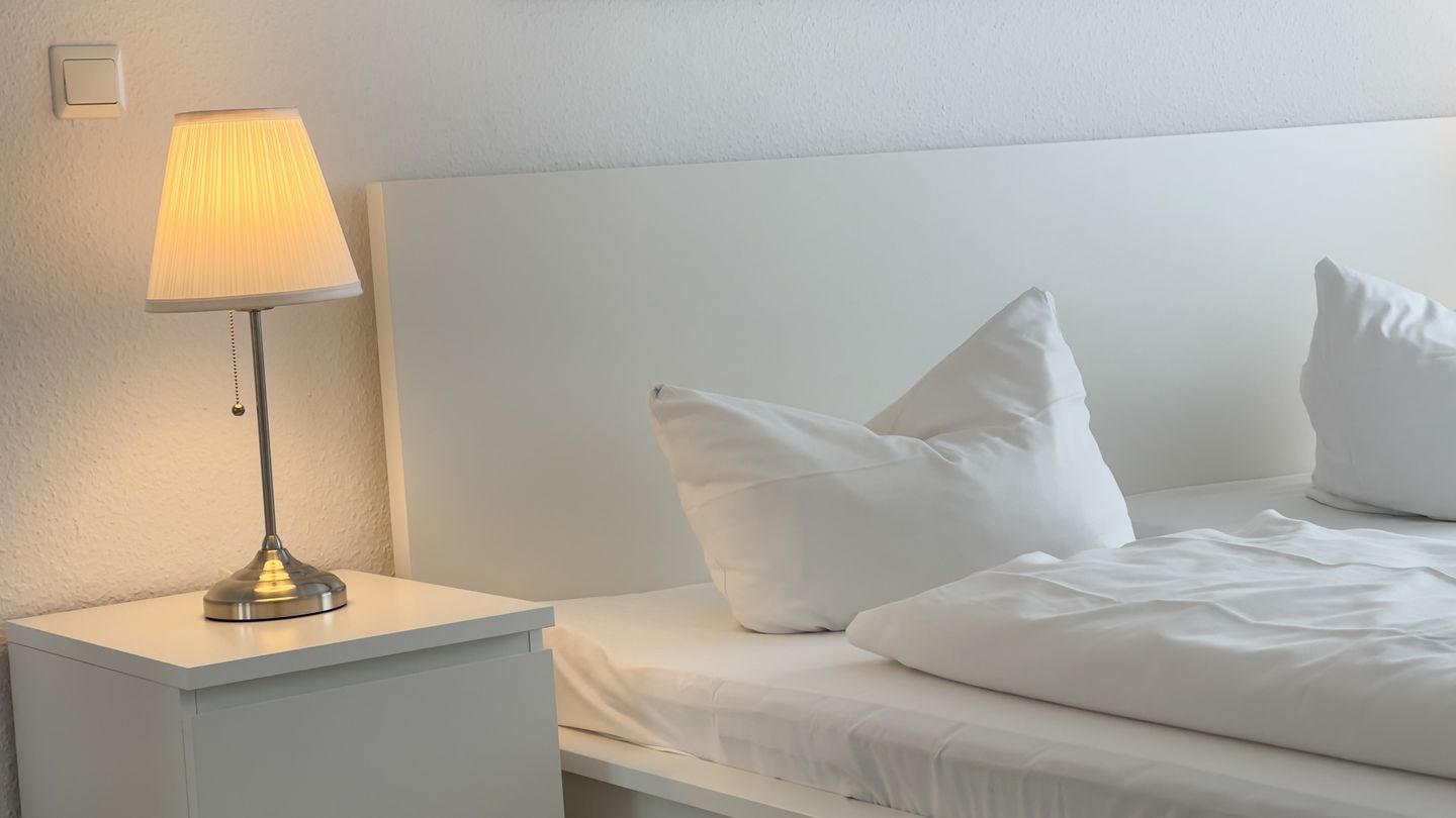 A neatly made bed with white bedding and pillows, a bedside lamp on a white nightstand, and a plain white wall.