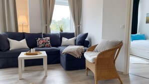 Un salon avec un canapé bleu foncé, une table basse blanche, un fauteuil en osier et, au fond, un autre canapé blanc.