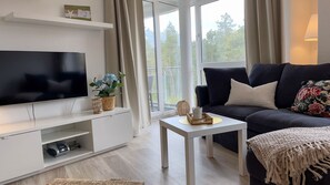 Ein Wohnzimmer mit Flachbildfernseher, einem dunkelgrauen Sofa, einem weißen Couchtisch und einem Fenster mit Blick auf Bäume.