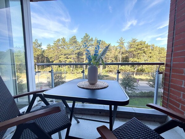 A balcony with a table and chairs, a potted plant, and a view of trees and a road.