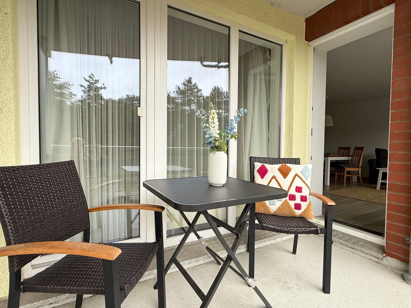 Eine Terrasse mit Tisch und Stühlen, eine Vase mit Blumen und Blick durch die Glastüren.