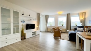 Ein modernes Wohnzimmer mit Flachbildfernseher, Essbereich mit Tisch und Stühlen, einer Couch und einem Schrank mit Glastüren.