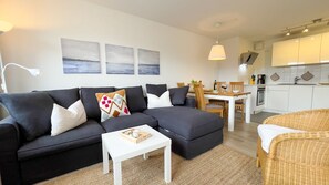 A modern living room with a sofa, a small table, and a wicker chair. There is a kitchen area with a dining table and chairs. - B2.2 - Spacious, modern apartment with balcony, only 200 m to the beach - House (Cuxhaven)