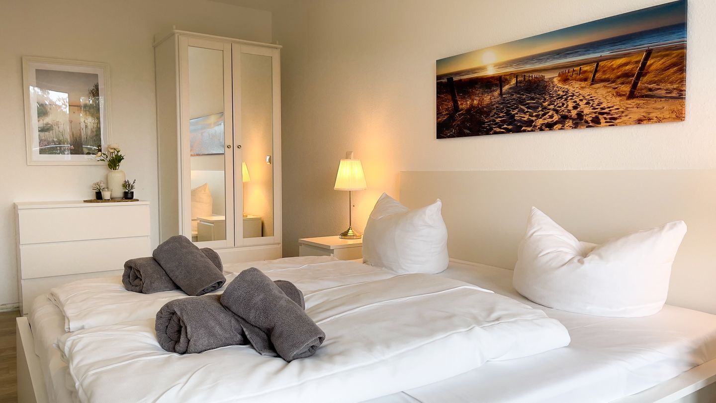 Ein ordentlich bezogenes Bett mit weißen und grauen Leinen, ein Nachttisch mit Lampe, ein Kleiderschrank und ein Gemälde mit einer Strandansicht an der Wand.