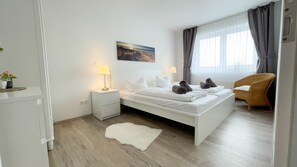 Un dormitorio moderno con una cama blanca, dos lámparas de noche, una alfombra blanca, una silla de mimbre y un cuadro en la pared.