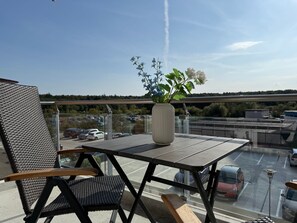 En balkon med bord, stole, en vase med blomster og udsigt til en parkeringsplads og bygninger.