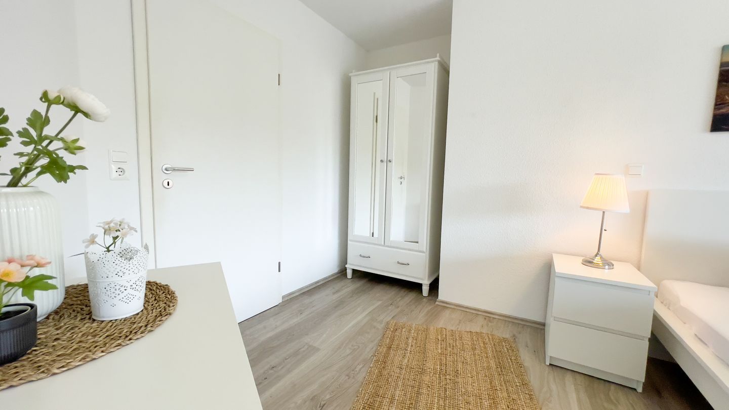 Une chambre avec un lit, une table de chevet, une lampe, une armoire et une plante posée sur une table.