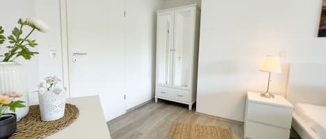A room with a bed, bedside table, lamp, wardrobe, and a plant on a table.
