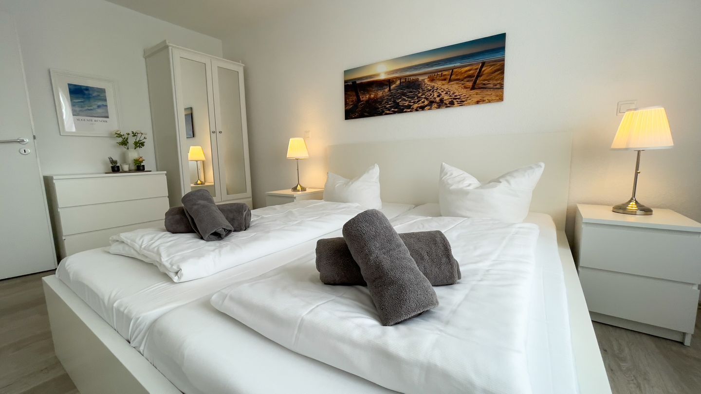 Een hotelkamer met twee bedden, wit bedgoed, grijze handdoeken, bedlampen, een schilderij aan de muur en een witte kledingkast.