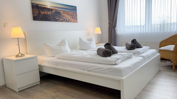 A double bed with white linens, two bedside lamps, a painting on the wall, and a wicker chair.
