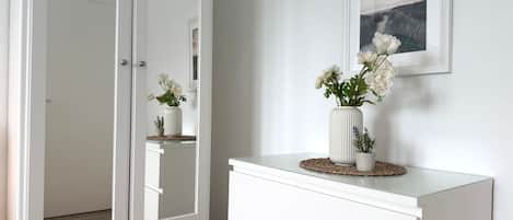 Un aparador blanco con cajones y un espejo, un jarrón con flores, y un cuadro en la pared.