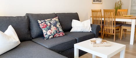 A grey sofa with floral and white cushions, a white coffee table with a candle and a wooden tray, and a wooden dining table with chairs in a room with light wooden flooring.
