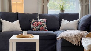 A dark grey sofa with floral and plain cushions, a white side table with a tray, and a view of greenery through a window. - A4.3 - Bright and inviting apartment with a partial ocean view, for 7 people (Cuxhaven)