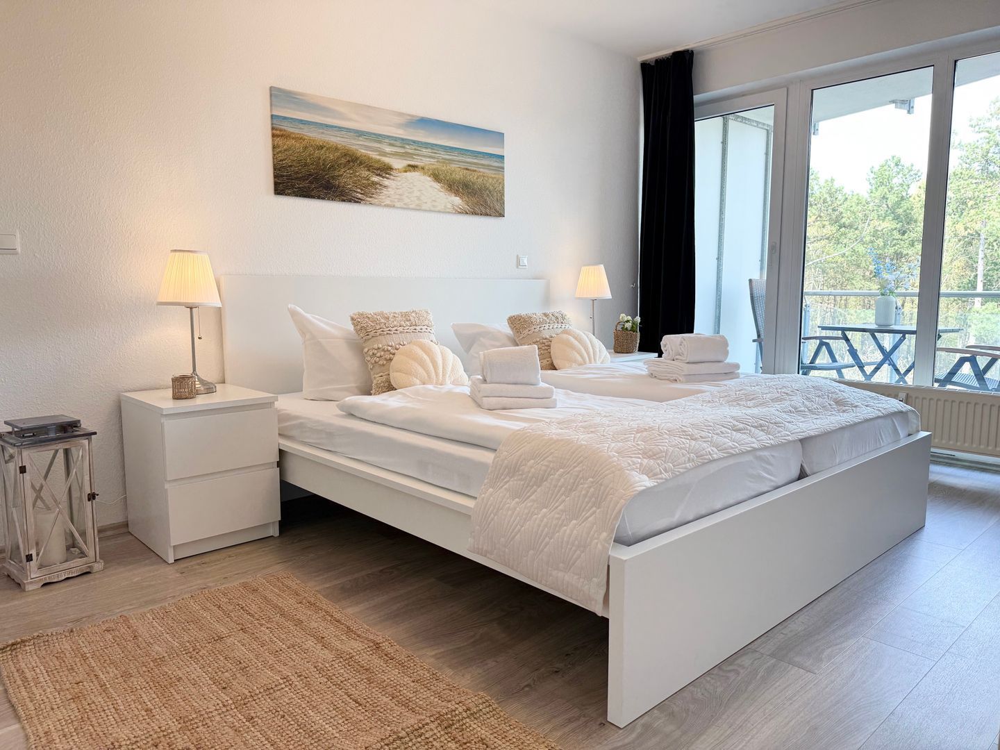 A bedroom with a large bed, two bedside tables, a nightstand, a lamp, a painting, and a view of the outdoors.