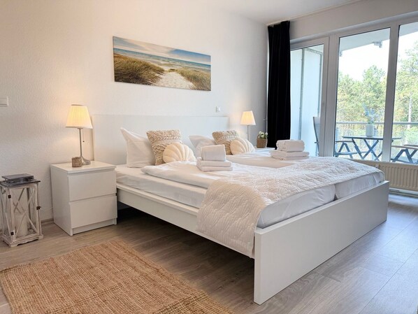 A bedroom with a large bed, two bedside tables, a nightstand, a lamp, a painting, and a view of the outdoors.