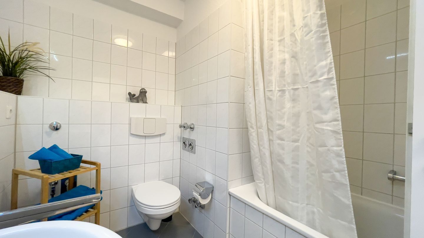 A bathroom with a toilet, a shower with a curtain, a sink, and a small shelf with a plant and blue towels.