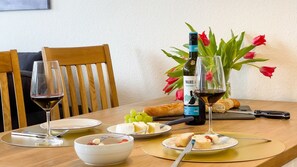 A dining table with a bottle of Malbec wine, two glasses of wine, a bowl of fruit, and plates of cheese and bread.