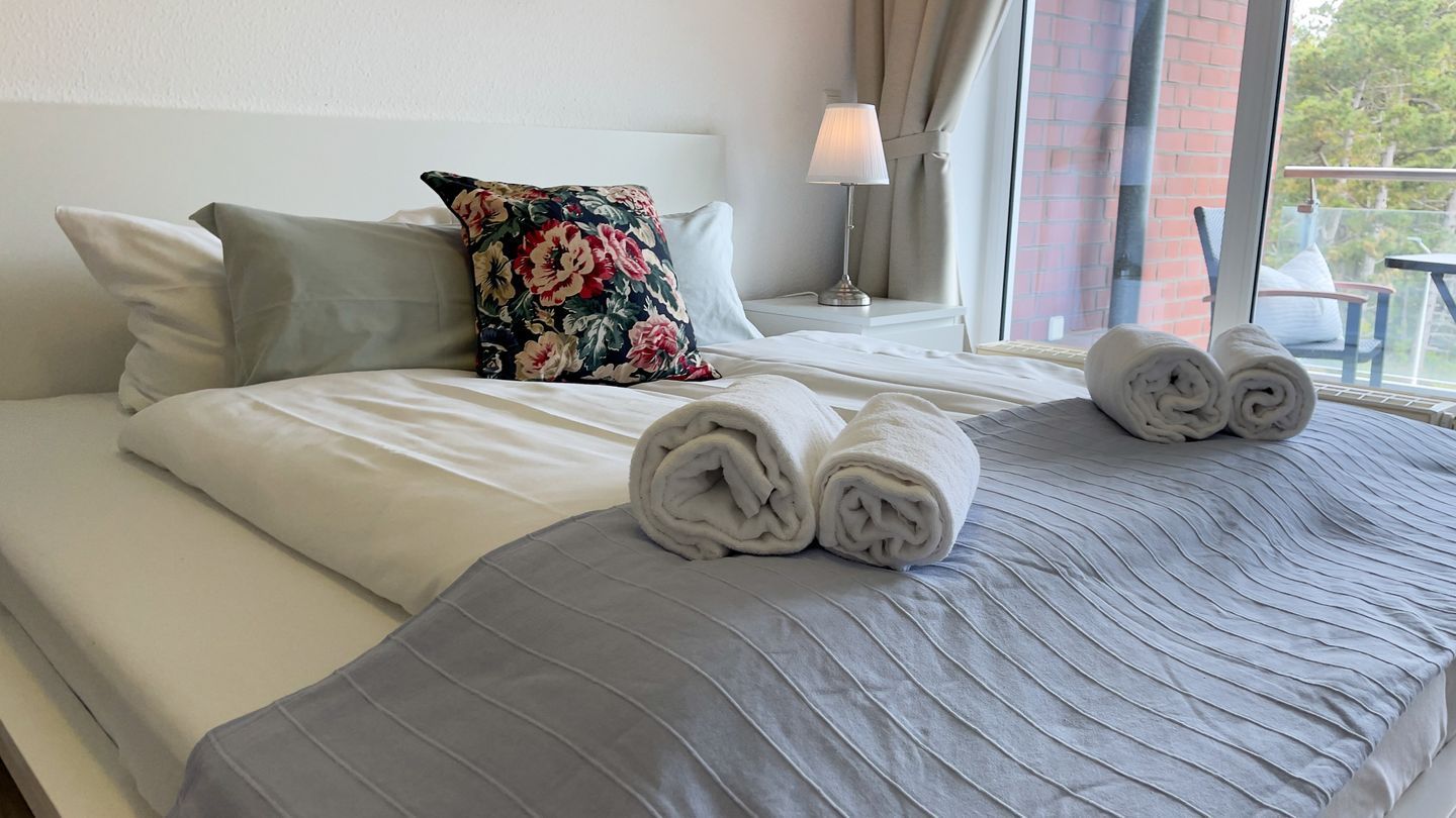 Un lit bien fait, avec des serviettes pliées, un coussin à motifs floraux et une lampe sur une table de chevet.
