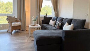 A living room with a sofa, a chair, a small table with a vase, and a window with curtains. - A3.3 - Bright vacation apartment, close to the beach for up to 7 people - House (Cuxhaven)