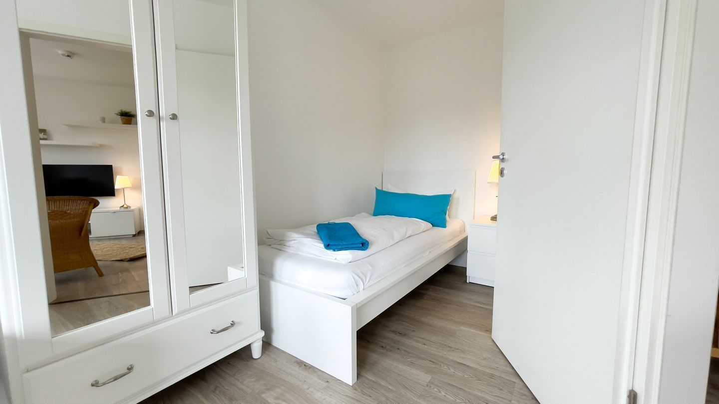 A small, white bedroom with a single bed, a nightstand, a mirror, and a wardrobe.