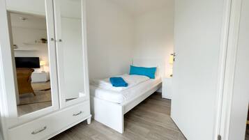 A small, white bedroom with a single bed, a nightstand, a mirror, and a wardrobe.