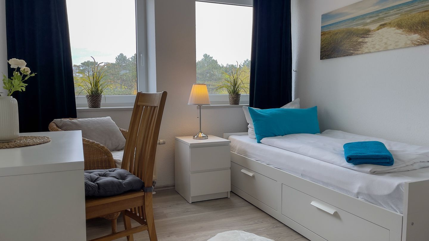 A bedroom with a bed, a chair, a nightstand, a lamp, and a window with curtains.