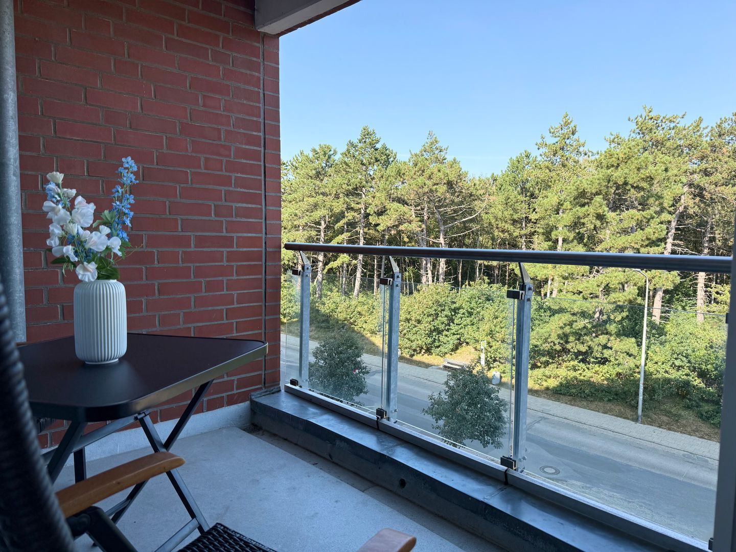 Un balcon donnant sur une route et des arbres, un vase blanc rempli de fleurs et un mur de briques.