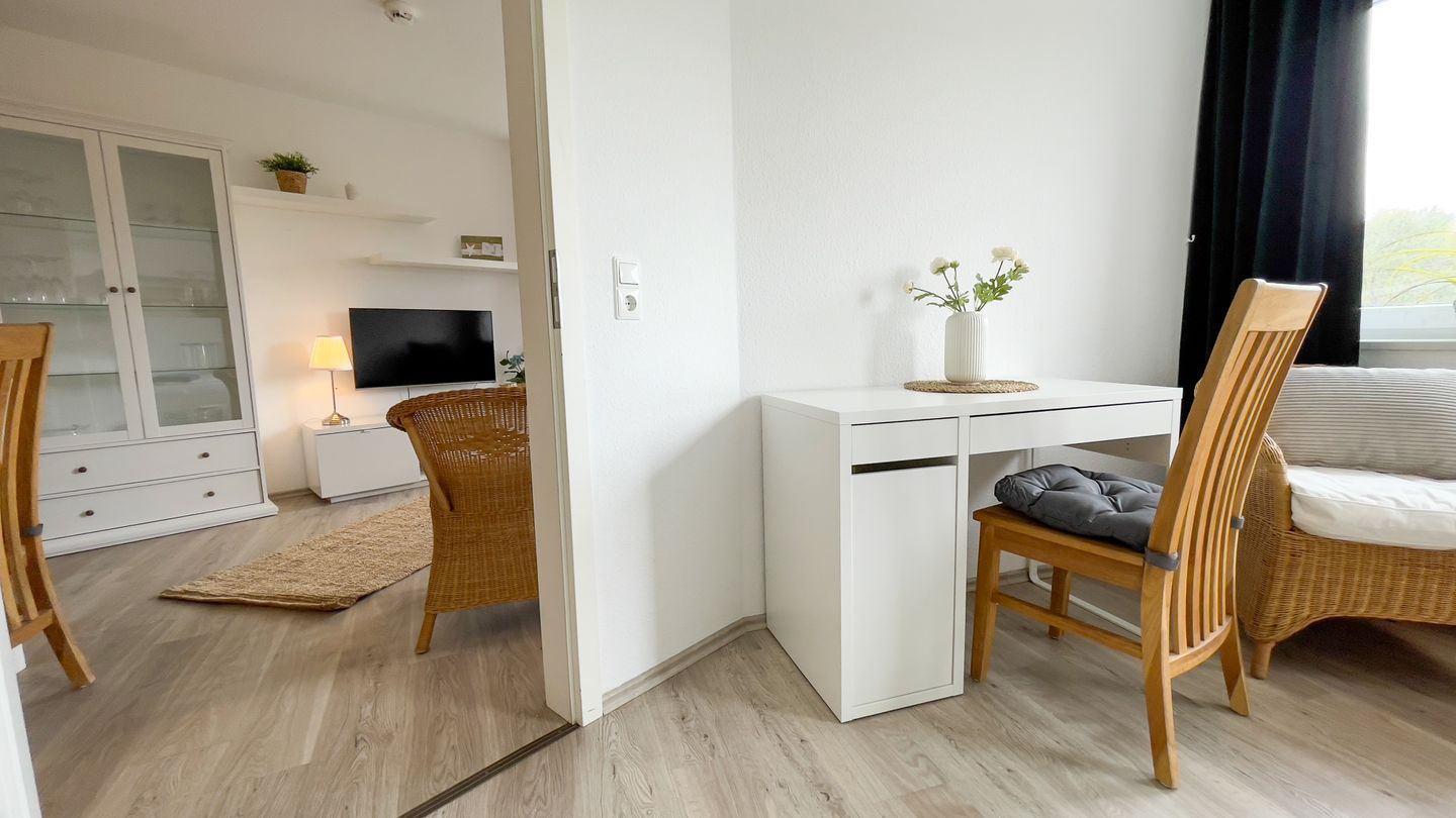 Una habitació amb escriptori blanc, cadires de veta, televisió, armari i finestra amb cortines.