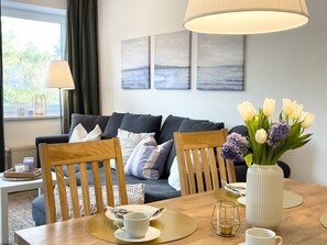 Un salon moderne avec un canapé, deux fauteuils en bois, une table à manger ornée d’un vase de fleurs et une lampe.