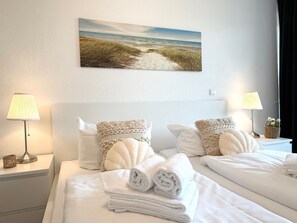 A hotel room with a bed, two bedside lamps, a painting of a beach, and a basket of flowers.