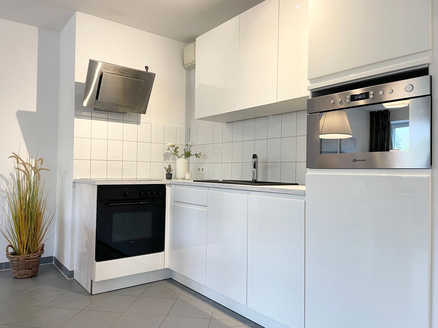 A modern kitchen with white cabinetry, a built-in oven, a stainless steel range hood, and a window above the sink.