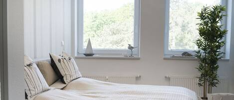 Un dormitorio con cama, almohadas, un radiador, una ventana con vistas y una planta.