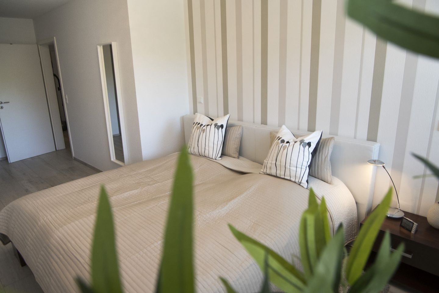 A bedroom with a bed, pillows, a bedside table, a lamp, and a plant.