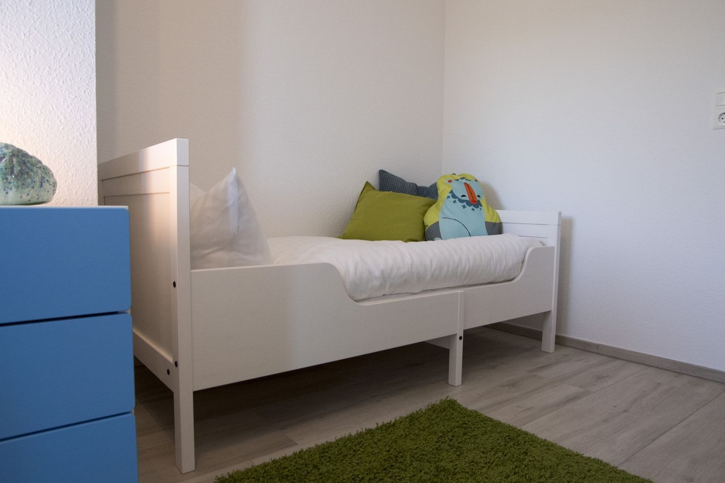 Ein weißes Bett mit Kissen und einem grünen Teppich in einem Raum mit einer blauen Kommode und einer weißen Wand.