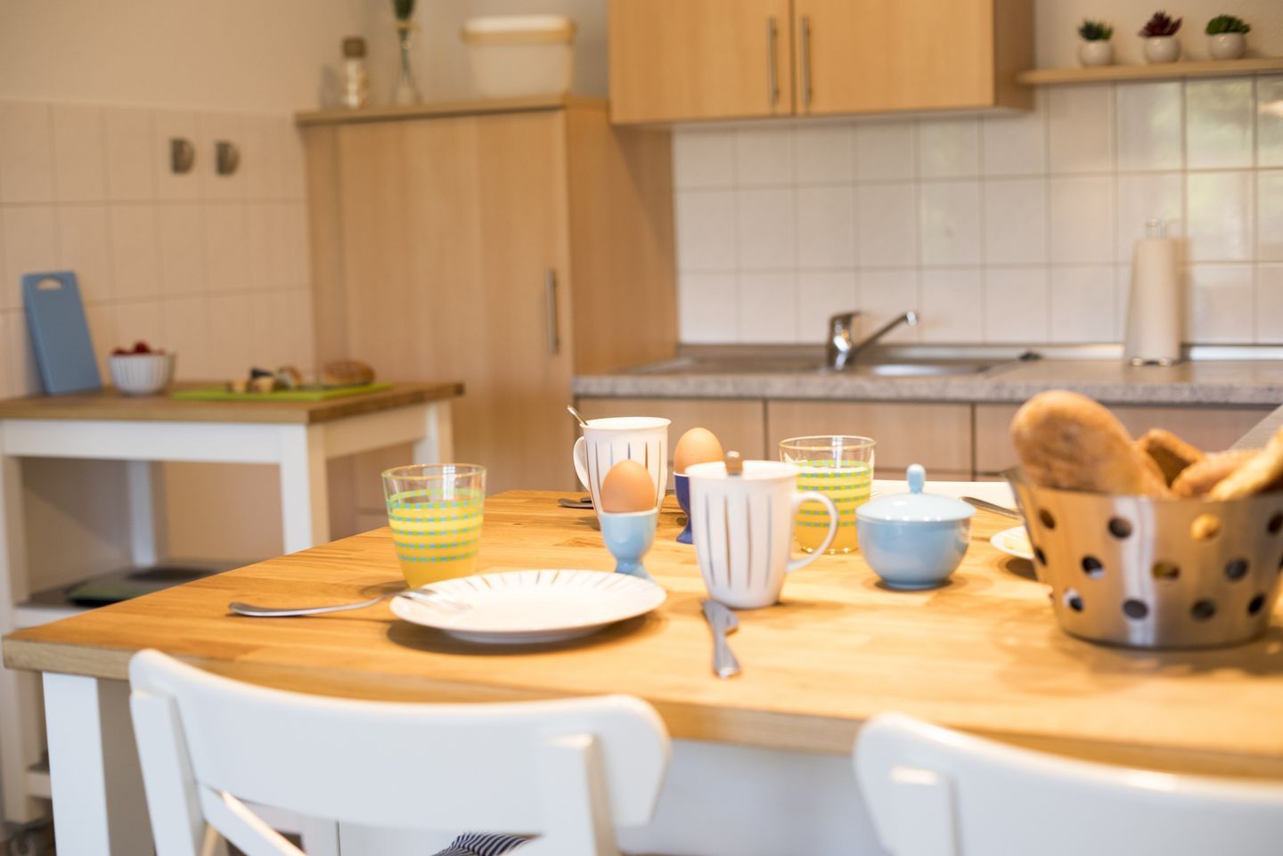 A kitchen with a wooden countertop, white chairs, and various kitchen items like a plate, cups, and a basket of bread.