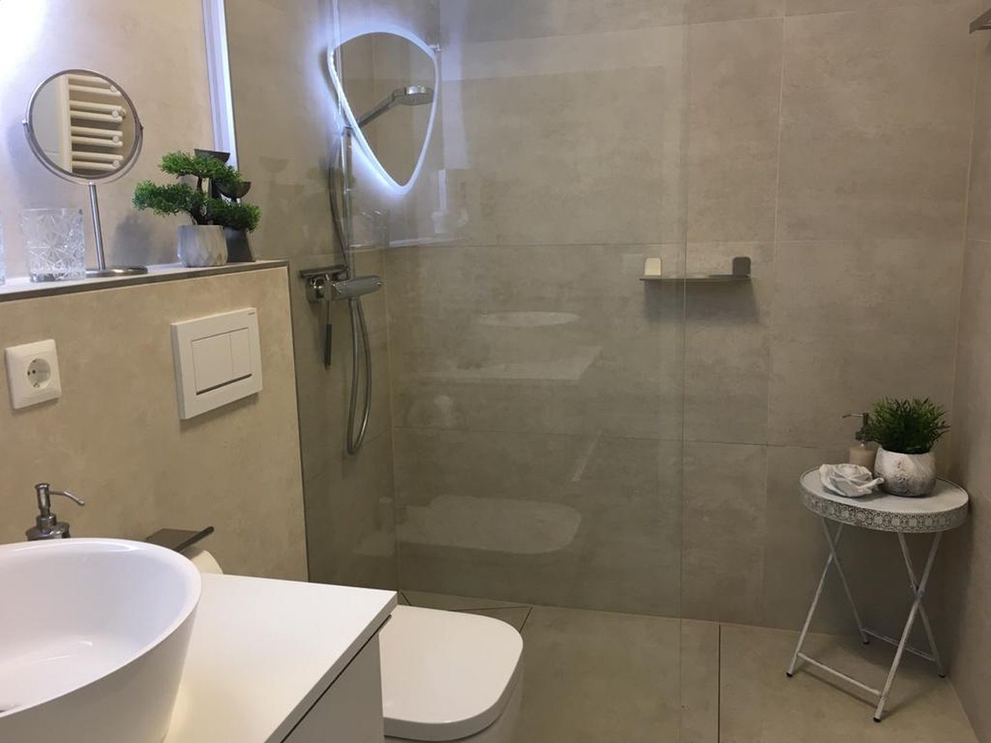 Un baño moderno con cabina de ducha de vidrio, un lavabo blanco y una mesita redonda con una planta.