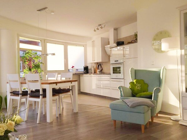 A modern kitchen with white cabinets, a dining table with chairs, a blue armchair, and a clock on the wall.