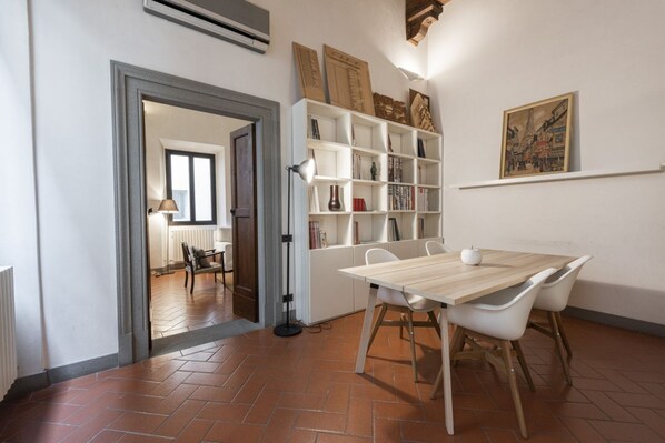 Un comedor con una mesa de madera y sillas blancas, una estantería y un cuadro en la pared.