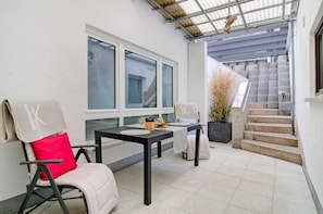En overdækket terrasse med bord og stole, en trappe og udsigt til gaden.