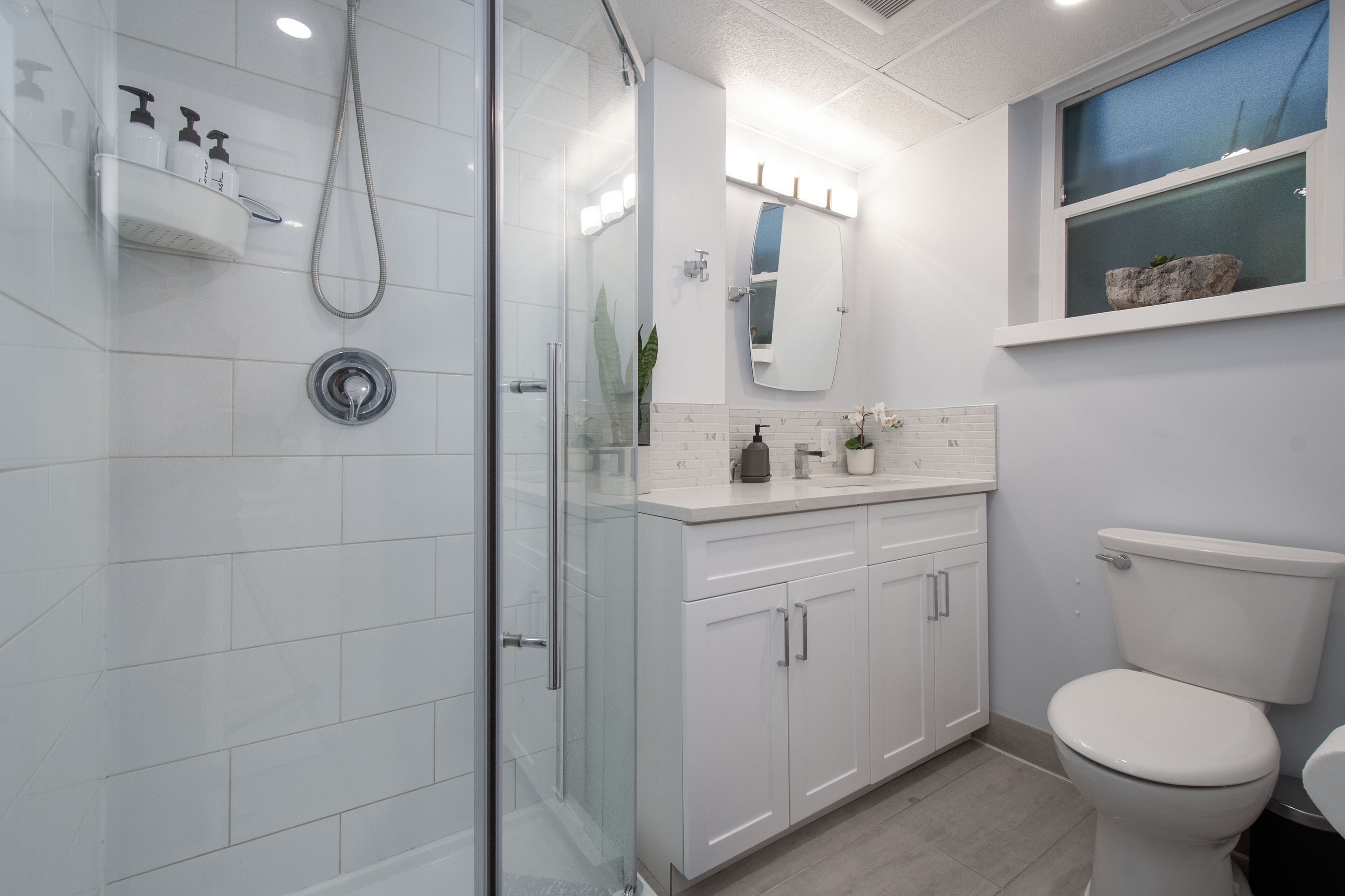 Une salle de bain moderne avec une douche à l’italienne en verre, des meubles blancs, des toilettes et une fenêtre agrémentée d’une décoration en pierre.