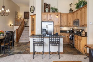 A kitchen with wooden cabinets, a stainless steel refrigerator, a microwave, and a stove. There are two bar stools and a dining table with chairs.