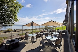 En udendørs terrasse med borde og stole, store parasoller og udsigt til vand.