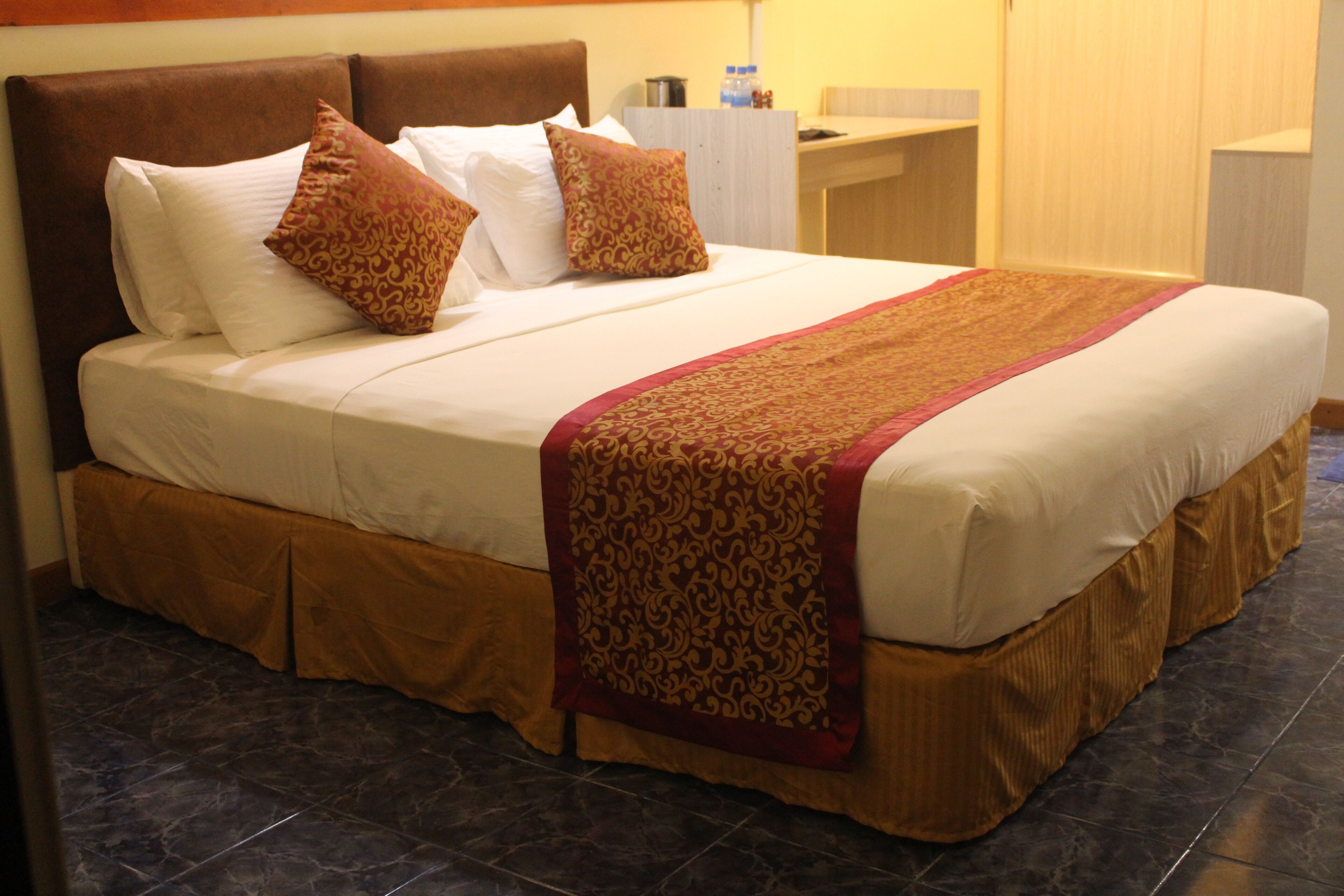 A neatly made bed with a patterned throw, a brown headboard, and a wooden nightstand with a bottle and a container.