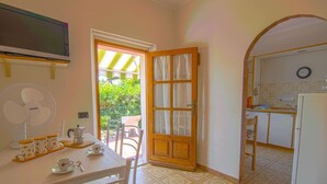 A dining area with a table set for two, a fan, and a view of the garden through a door. - Le Mimose - Two-Room Apartment (Campo nell'Elba)