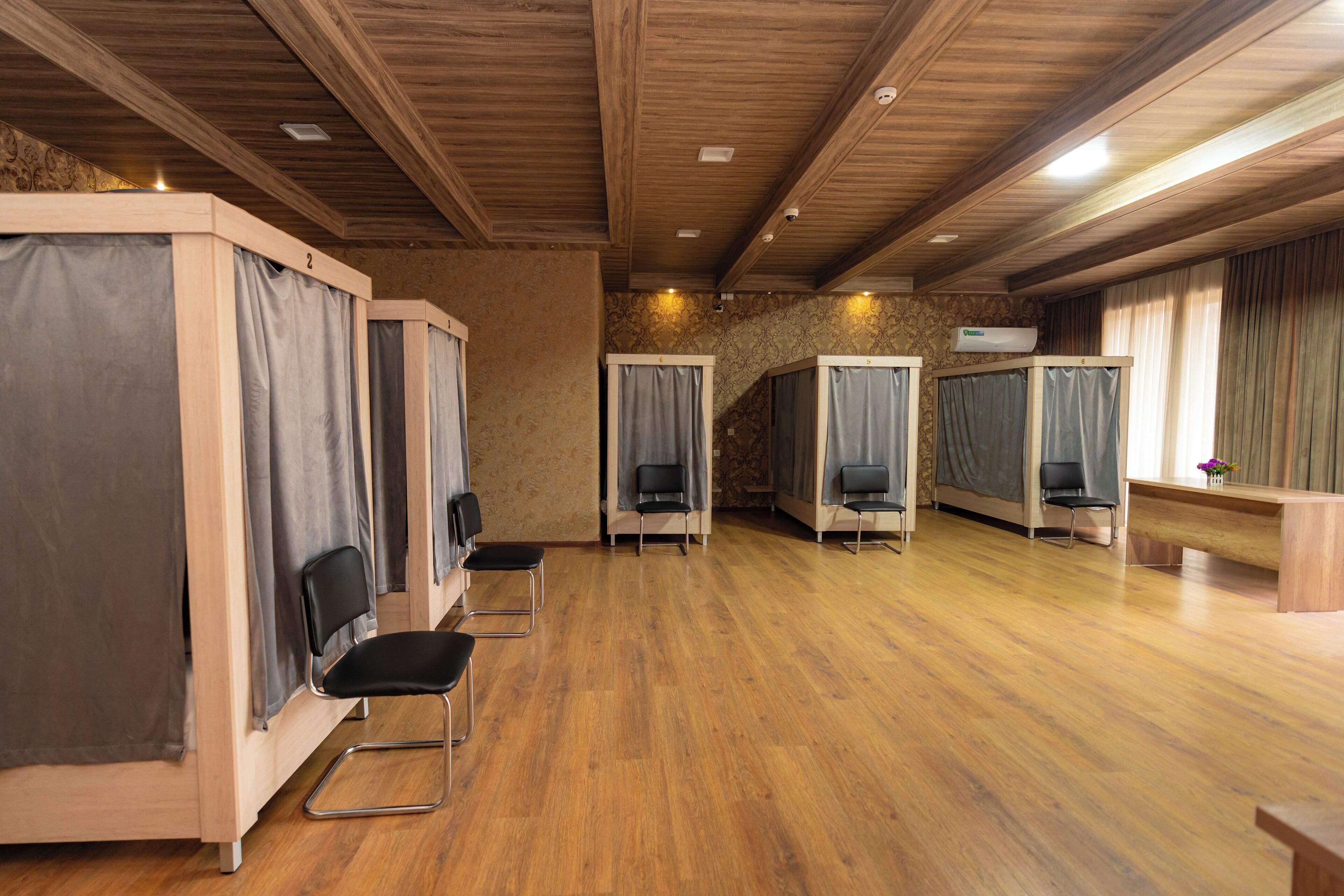 A room with wooden floors, wooden ceiling beams, and a reception desk. There are four individual seating areas with curtains and chairs.