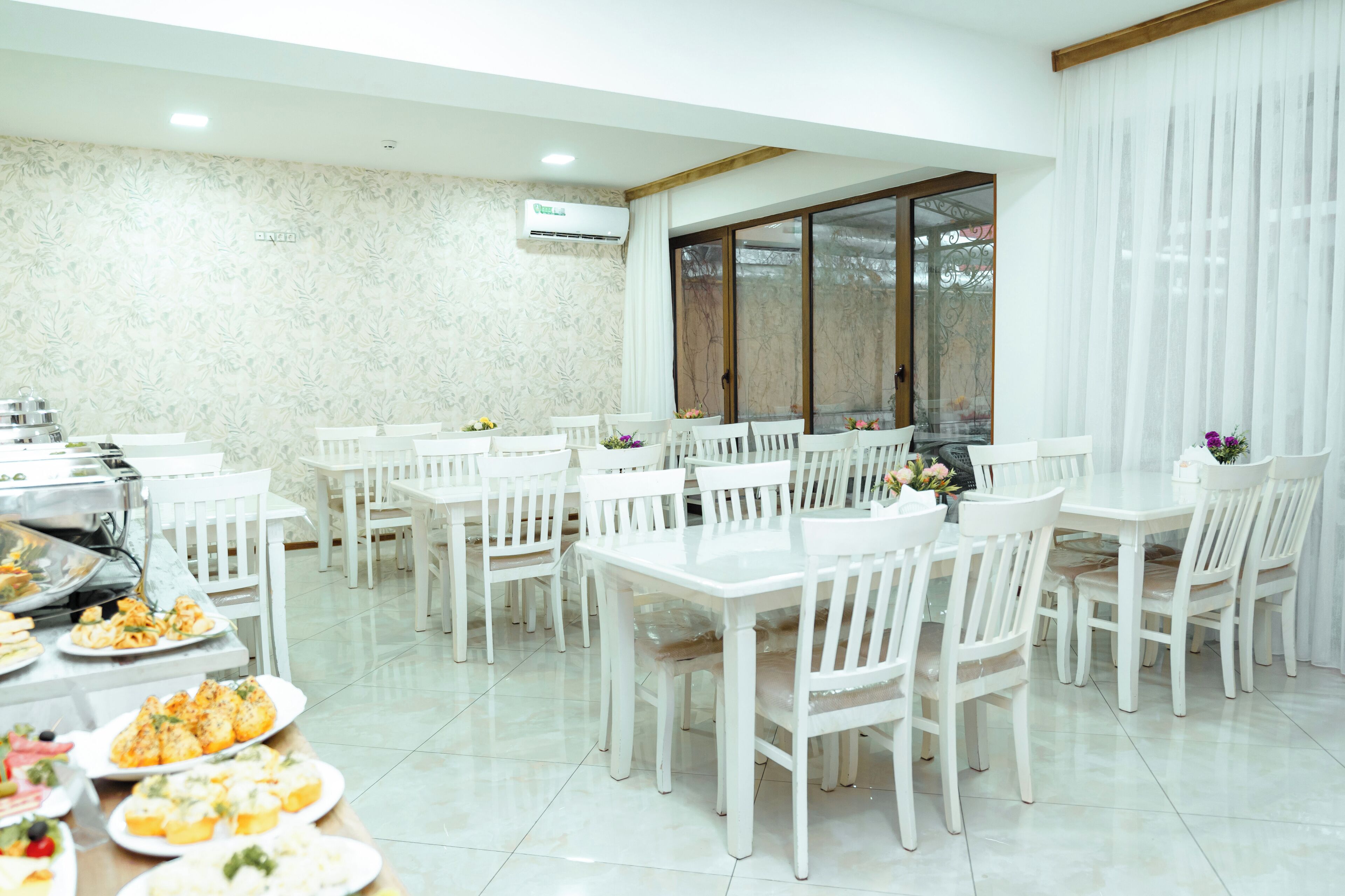 A dining area with white tables and chairs, a buffet with various dishes, and a large window with curtains.
